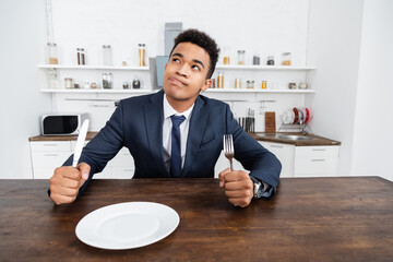 dreamy african american man holding cutlery while thinking and sticking out tongue.