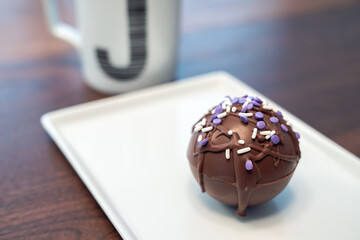 One hot cocoa bomb ball or sphere covered in drizzled brown chocolate and purple and white sprinkles set on a white serving plate with monogram J coffee mug nearby making a delicious winter treat.