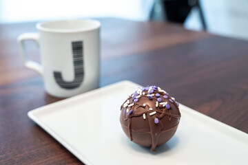 One hot cocoa bomb ball or sphere covered in drizzled brown chocolate and purple and white sprinkles set on a white serving plate with monogram J coffee mug nearby making a delicious winter treat.