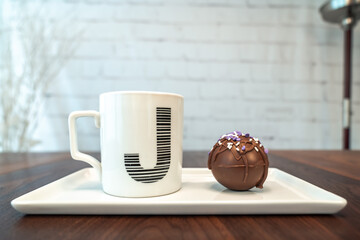 Side view of a hot cocoa or chocolate bomb covered in drizzled brown chocolate and purple and white sprinkles and letter J monogram coffee mug on plate and wood table top with white brick background.