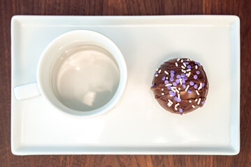A top view food photograph of a hot cocoa bomb covered in drizzled brown chocolate and purple and white sprinkles set on a white serving plate next to a coffee mug or cup on a dark wood table top.
