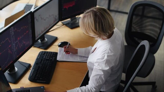Writing information on the document. Female stockbroker in formal clothes works in the office with financial market and graphs on monitors.