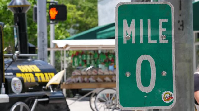 The mile marker 0 sign at the end of US Highway 1 in Key West Florida and a tour train drives by in the background, slow motion.