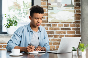 Obraz premium surprised african american man holding pen near notebook and looking at laptop.