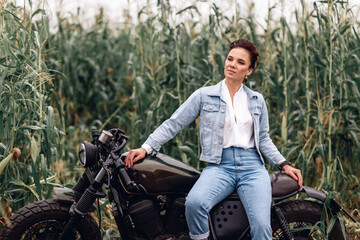 Lovely beautiful young woman wearing denim clothes and sitting on a large black retro motorcycle against a backdrop of grass and greenery. Concert of speed