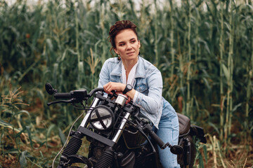 Portrait of an adult pretty stylish woman wearing denim casual clothes and sitting and posing on a black retro motorcycle. Green background. Extreme concept