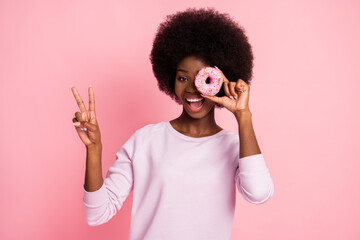 Portrait of nice cheerful wavy-haired girl closing eye with fresh yummy donut showing v-sign isolated over pink pastel color background