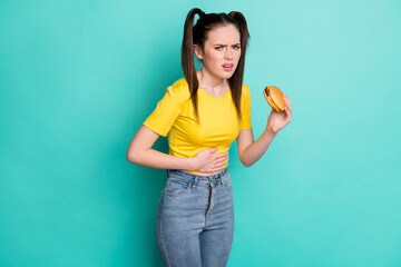 Photo of sad unwell young woman dressed yellow t-shirt holding burger overeating arm hand stomach...