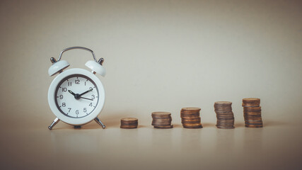 white analog clock with blurred pile of coins on brown background for business and finance concept , time for savings money concept, banking and business idea. vintage tone
