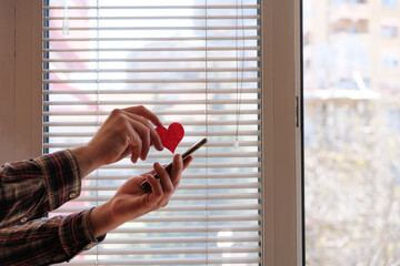 Red heart and mobile phone in hand.  Sending love message concept. Indoor shot near the window at day. Modern hipster lifestyle