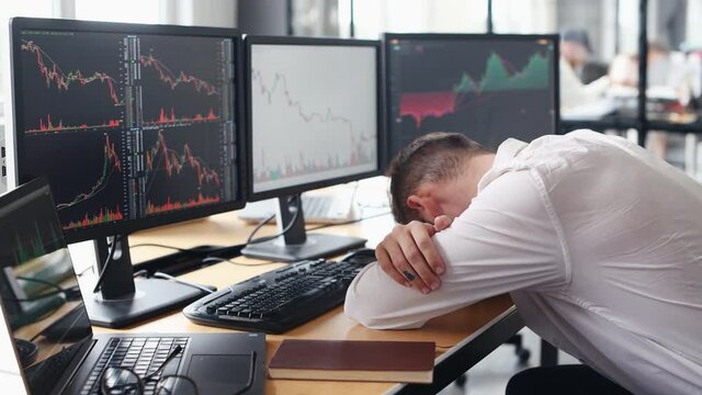 Tired male stockbroker in formal clothes leaning by the table and sleeps in the office with financial market and graphs on monitors.