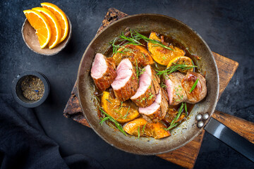 Traditional fried pork filet medaillons in with orange slices and herbs offered as top view in a rustic wrought iron skillet - obrazy, fototapety, plakaty