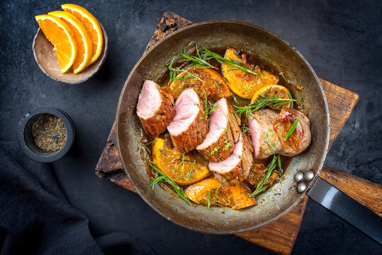 Traditional fried pork filet medaillons in with orange slices and herbs offered as top view in a rustic wrought iron skillet