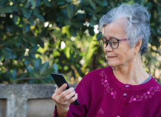 Portrait of a senior woman wearing glasses holding and looking at a smartphone while standing in a garden. Space for text. Concept of old people and technology