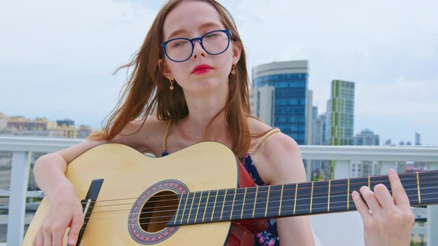 A young woman plays the guitar on the roof of a multistory building in the city