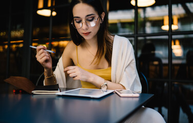 Caucasian blogger in glasses installing gadget application for online messaging and chatting, millennial hipster girl 20 years old booking tickets via touch pad during web networking in street cafe