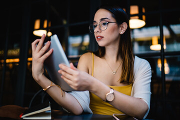 Millennial female blogger in spectacles holding modern digital tablet reading book online during break, attractive female freelancer share publication and multimedia content via touch pad with 4g