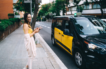 Cheerful hipster girl in spectacles holding cellphone gadget using for online booking taxi cab looking for transport near city road with traffic, happy millennial generation Z smiling outdoors