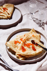 Traditional Russian crepes or pancakes with red caviar on gray plates and glasses of ice vodka over greige linen tablecloth. Pancake week or Shrovetide