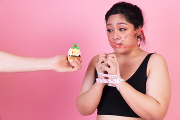 Diet restricts, weight loss, eating disorder concept. Obese overweight fat woman with measure tape cover her mouth while hands of man holding cupcake her thinking eat it over isolated pink background.