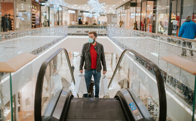 A handsome man in a leather jacket with a medical protective mask on his face, climbs the escalator in the mall. Covid-19