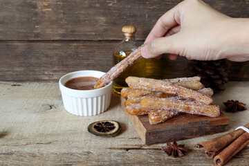 right hand dipping the homemade Spanish caramel churros into chocolate dipping sauce pasty on the wooden table with cinnamon sticks on vintage wood kitchen wall background during christmas party