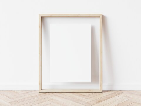 Blank vertically oriented rectangular picture frame with light wood border standing on wooden floor leaning on white wall. 3D Illustration.