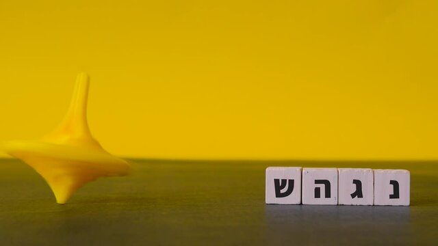 Spinning yellow top Hanukkah careers about beside the four hebrew letters nun, gimel, heh and shin shown in a static shot against a two-tone background.
