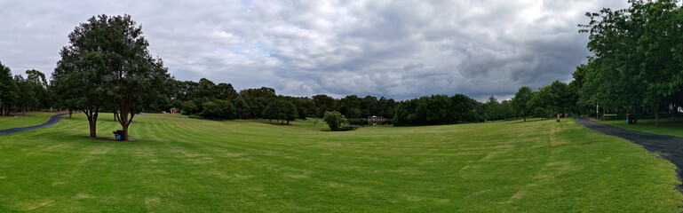 Fototapeta na wymiar Beautiful panoramic morning view of a park with green grass, tall trees and dark cloudy sky, Fagan park, Galston, Sydney, New South Wales, Australia 