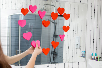 Hands of young girlwoman in the bathroom glue valentines red hearts on mirror in bathroom....