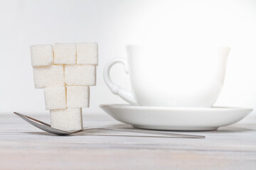 Lots of sugar lumps on a spoon near a white mug on a wooden white table.