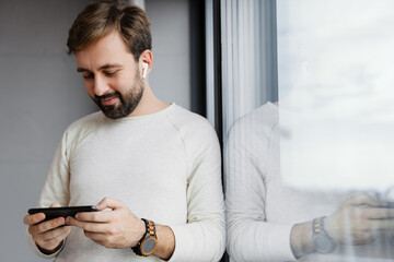 Handsome pleased man using cellphone while leaning on window glass