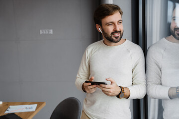 Handsome happy man in earphones using cellphone while standing