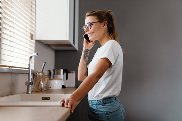 Beautiful happy girl smiling while talking on mobile phone at home kitchen