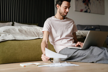 Focused man working with papers and laptop while sitting on floor