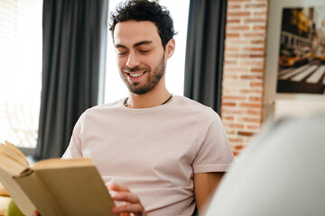 Happy unshaven man smiling and reading book while sitting on couch