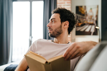 Focused unshaven man reading book while sitting on couch at home