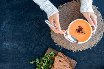 A woman in a white sweater eats pumpkin soup with fried bacon with bread. Pumpkin cream soup in a white plate on a napkin on a black background. Top view.