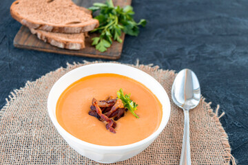 Pumpkin soup with fried bacon in white plate on a black background with bread and parsley. Top view.