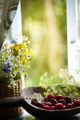Ripe red cherries and wildflowers on an old wooden window overlooking the garden. Vintage photo with summer mood. Soft selective focus.