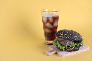 black hamburger on wooden cutting desk and a glass of ice cola isolated on yellow background. Unhealthy food concept
