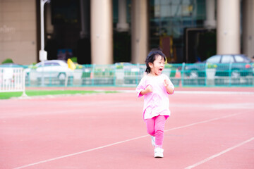 Fototapeta premium Active girl running have fun on red track in the stadium. Children were happy, smiling and laughing. Child exercise. Cute kid wearing a pink dress is 3-4 years old.