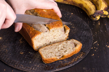 Female hands cut a traditional American dish with a knife - gluten-free and dairy-free banana bread on a black concrete background.