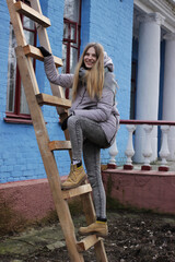 Obraz na płótnie Canvas A cheerful blonde girl in winter clothes climbed the wooden stairs near the house.
