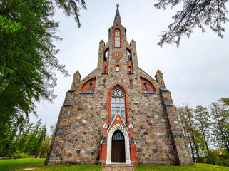 Latvia - The Cesvaine Lutheran Church, a building of colored stones with a clock, against a blue sky, surrounded by green trees.