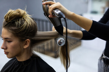 Naklejka premium Stylist making hairstyle using hair dryer, blowing on wet customer hair at beauty salon