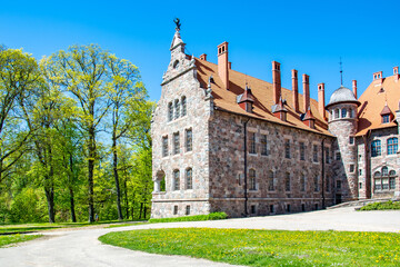 Cesvaine, Latvia - 05.23.2020: Cesvaine castle - a manor house of the late 19th century, a building of stones of different colors with a brown tiled roof, green trees and blue sky.