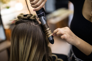 Young woman hairdresser making curls at blond hair with curling irons at luxury barber salon. Stylist and beauty