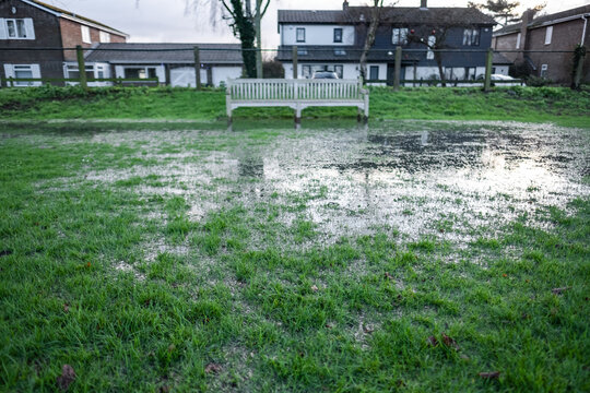 Shallow focus of grass seen on a public park having been subjected to heaving storms. The area is now waterlogged and a distant park bench is visible.