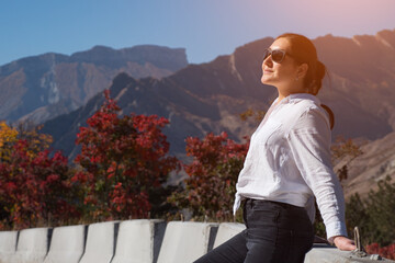 Fototapeta na wymiar Young woman in sunglasses poses near concrete road barriers against orange yellow autumn trees and rocky hills silhouettes under blue sky